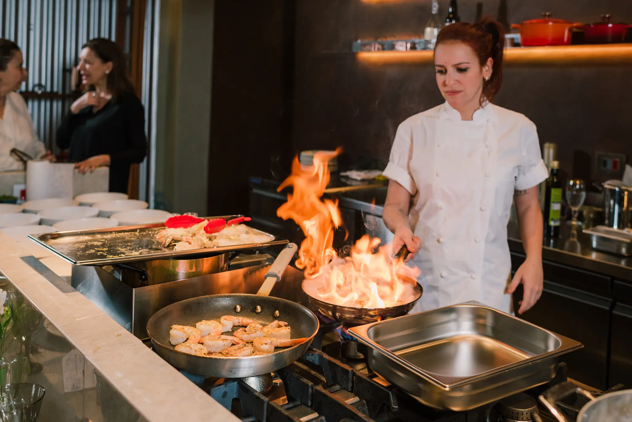 Chef Sofia Maria flambando durante uma aula de culinária expositiva, com alunas ao redor do fogão